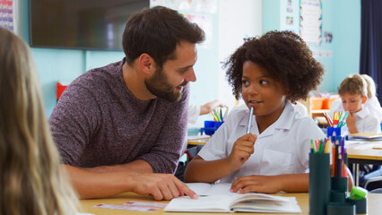 Male Elementary School Teacher Giving Female Pupil Wearing Uniform One To One Support In Classroom 