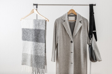 Light grey wool coat or jacket,  alpaca scarf and black leather bag on clothes hanger against white wall in home hallway. Autumn and winter wool  female clothing. Trendy monochrome fashion look.