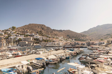 The vibrant harbor of Hydra Island showcases numerous boats docked in calm waters, surrounded by quaint buildings and steep hills under a clear blue sky on a sunlit day.