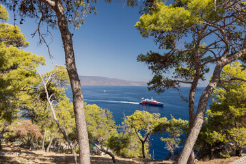 A colorful boat glides through the clear blue waters off the coast of Hydra, Greece, surrounded by vibrant green trees under a bright sunny sky.