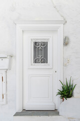 The entrance features a beautifully crafted white door with decorative wrought iron, surrounded by whitewashed walls and a potted plant, typical of Hydra's architecture.