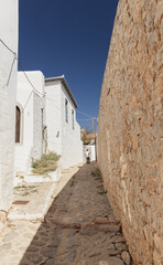 A picturesque alleyway on the Greek island of Hydra, flanked by traditional whitewashed houses. A solitary figure walks along the cobblestone path, enjoying the serene atmosphere.