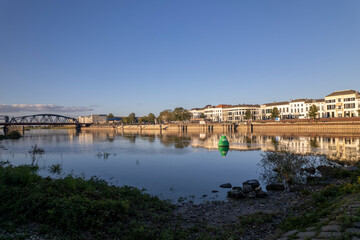 Obraz premium Floodplain view of Zutphen countenance against a clear blue vibrant colourful sky with quay visible of river IJssel in low water level, historic tower town reflecting in the waterway