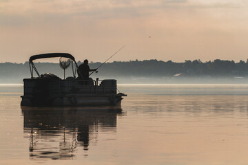 Silhouette of a lone man casting his rod while fishing on his pontoon boat in the early morning mist.