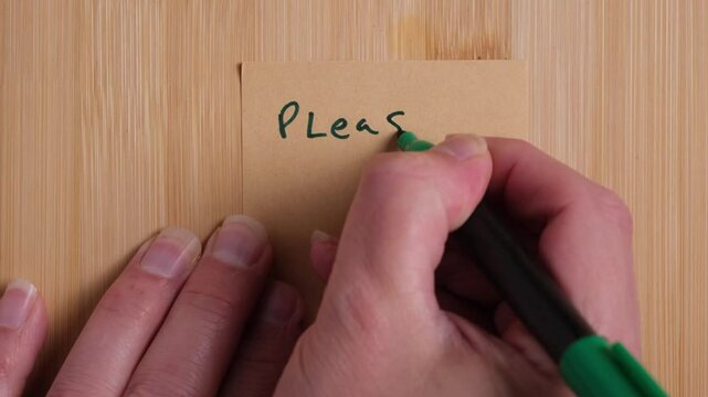A person is writing on a recycled paper sticky note on a sustainable source wood background. The person is writing Please Recycle. Concept, sustainability, green, climate.