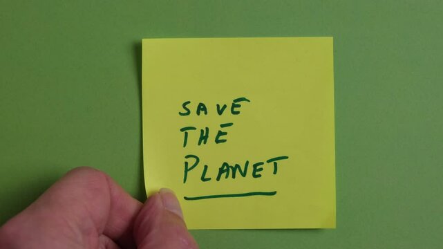 A person is placing a sticky note on a green background. Written on the sticky note are the words Save The Planet. Concept, sustainability, ecology, climate. Slow motion.