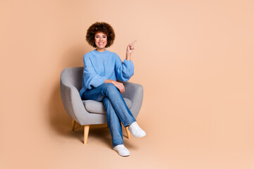 Full size photo of cute girl dressed knit sweater jeans sit on armchair directing at promo empty space isolated on pastel color background