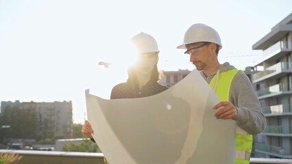 Two architects wearing safety hard hats are holding blueprint and discussing something on construction site at sunrise. Architect and teamwork concept