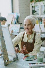 Vertical shot of silver haired woman smiling widely while doing strokes of paint brush on white...