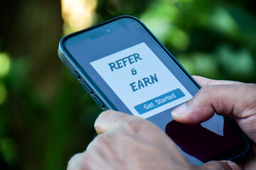 A man mobile phone displays the Refer and Earn program on the screen. Customer rewards, referral program to make money concept. Digital Marketing strategy for increasing sales. Selective focus.