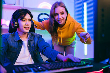 A young man plays video games while a woman enthusiastically guides him, surrounded by colorful LED...
