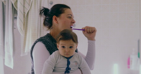 Mother multitasking in the bathroom, brushing her teeth while holding her baby. The baby gazes...