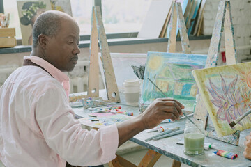 Senior African American man sitting at dirty desk with painting tools and wetting paintbrush in jar...
