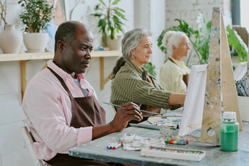 Small group of biracial retired people painting scenery on canvases during art therapy together