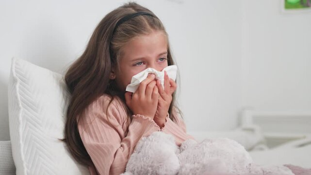 Sick girl sneezing snot into tissue paper in bedroom at home. Female child blowing runny nose is suffering from cold and flu 