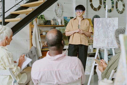 Back view shot of biracial senior student listening young adult female teacher giving drawing lesson in white studio