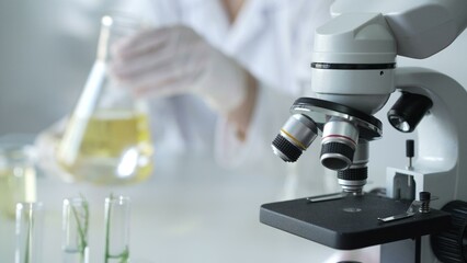 Microscope is near a scientist, wearing a lab coat and white protective gloves, and holding beaker with a yellow oily liquid in laboratory setting, close up. Medicine and science concept