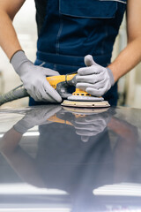 Vertical cropped shot of unrecognizable mechanic male in protective gloves polishing surface of hood of car using orbital polisher in repair shop. Concept of professional car repair, maintenance work.