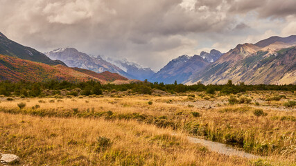 This stunning landscape, located near El Chaltén in Patagonia, Argentina, features autumn colors, mountainous terrain, and a serene stream winding through the grassy fields below the Andes.