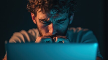 A focused young man gazes intently at a laptop screen, showcasing deep concentration amidst a darkened environment.