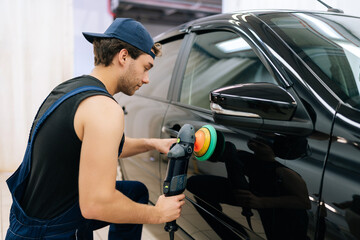 Rear view of muscular mechanic worker polishing door of luxury car in auto repair shop using with orbital polisher. Expert car paint technician using car polisher to polish car with refinement.