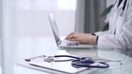 Fototapeta premium Tablet computer and stethoscope are lying on the glass table while doctor woman is using laptop computer on the glass table. Medicine concept