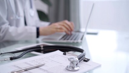 Fototapeta premium Clipboard with medical records and stethoscope are lying on the glass table while doctor woman is using tablet computer on the glass table. Medicine concept