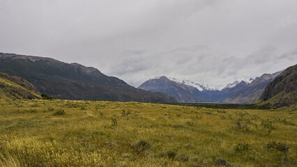 This serene valley scene is near El Chaltén, Patagonia, Argentina, showcasing rolling grasslands framed by rugged mountains and distant snow-capped peaks, creating a tranquil atmosphere.