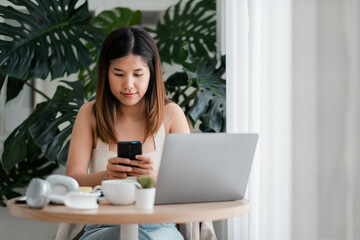 Fototapeta premium A young woman sits at a table using her smartphone and laptop, surrounded by green plants in a modern home office setting.