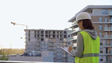 Woman engineer with a hard hat and safety vest is writing on a clipboard while inspecting a construction site at sunset