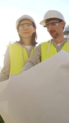 Man and woman architects or engineers wearing safety hard hats and vests holding blueprint and...