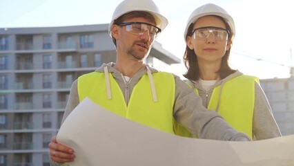 Architects or engineers wearing safety helmets and vests holding blueprint and discussing something...