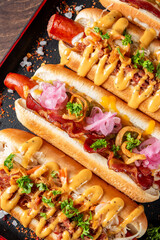 Close-up of three hot dogs with various toppings, including mustard, ketchup, onions, and herbs, on a dark tray. Perfect for illustrating fast food, American cuisine, or street food concepts.
