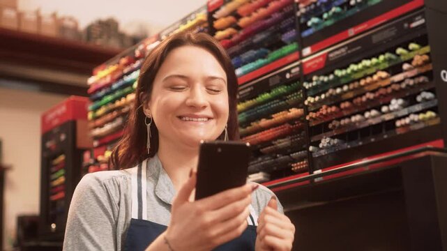 Excited female store employee celebrating success while checking phone. Happy young woman enthusiastically reacting to good news in vibrant art supplies store