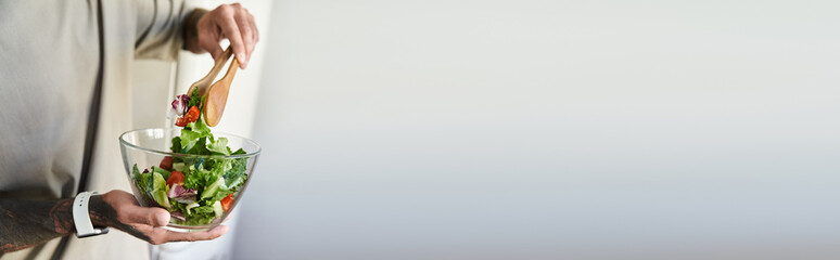 A young man joyfully prepares a vibrant salad, showcasing fresh ingredients in his cozy kitchen.