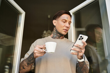 A handsome young man stands on his balcony, sipping coffee and browsing his phone, relaxed at home.