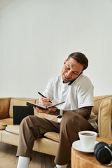 A young man sits comfortably at home, engaging in phone conversations and taking notes.