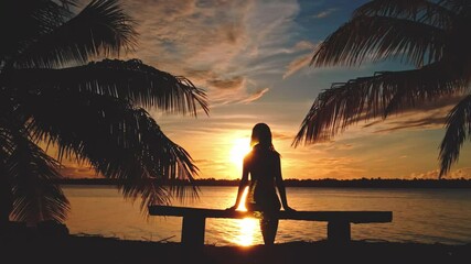 Silhouette of a woman sitting on a bench by the sea, enjoying a beautiful sunset over a tropical island with palm trees