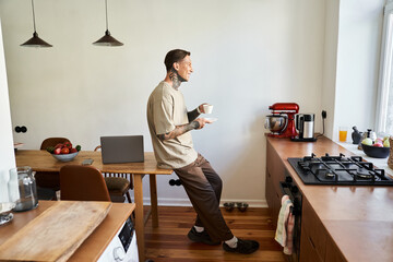 Relaxing at home, a young man sips coffee and focuses on his tasks in a sunlit kitchen.