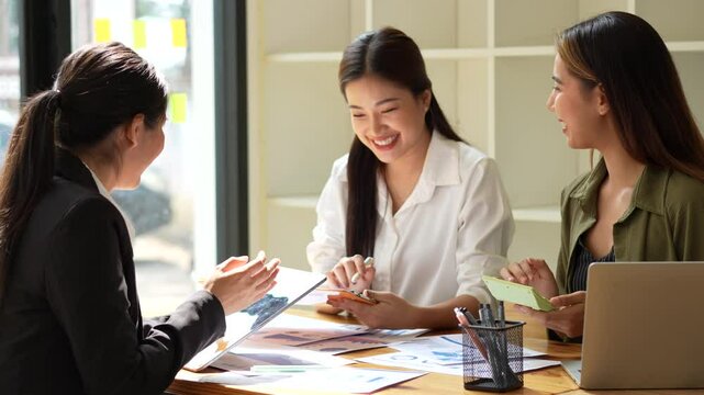Three women in modern workspace engage in lively discussion, showcasing teamwork and collaboration through creativity while using digital devices.
