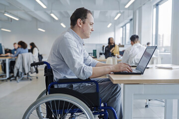 Disabled man sits in a wheelchair and works in an office on a laptop.