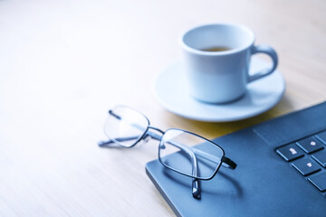 Part of a black laptop, glasses and a cup of coffee on a wooden desk in an office, concept for business, study, writing or journalism, copy space, selected focus