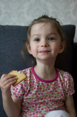 Naklejka premium A small child or girl or baby sits on a sofa or armchair, holds a cookie in her hands, smiles and eats a cookie. There are cookie crumbs on the child's face