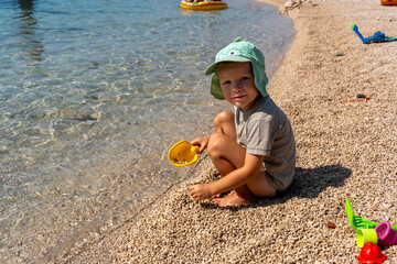 Young boy playing with sand toys at the beach, wearing a cute green sun hat on a sunny day. Concept of summer fun, childhood joy, and outdoor activities by the water.