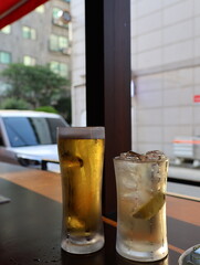 Two drinks on a table, one is a beer and the other is a cocktail. The beer is in a tall glass and the cocktail is in a shorter glass. The scene takes place in a bar or restaurant