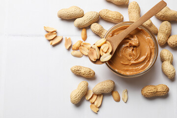 Tasty peanut butter in bowl and groundnuts on white tiled table, flat lay. Space for text