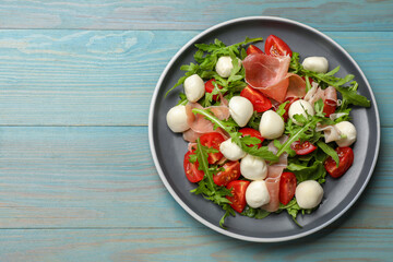 Tasty salad with arugula, mozzarella cheese, tomatoes and jamon on light blue wooden table, top view. Space for text