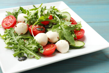 Tasty salad with arugula, mozzarella cheese and vegetables on light blue wooden table, closeup