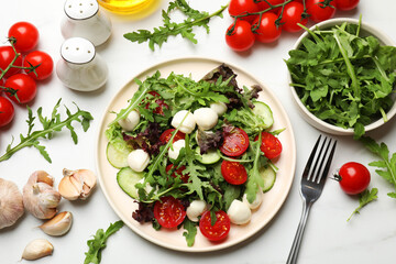 Tasty salad with arugula, lettuce, mozzarella cheese, vegetables and fork on white table, flat lay