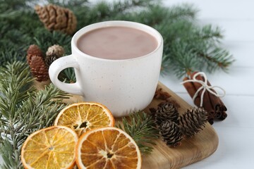 Yummy cocoa in cup, dried orange slices, cinnamon sticks and fir tree branches on white wooden table, closeup